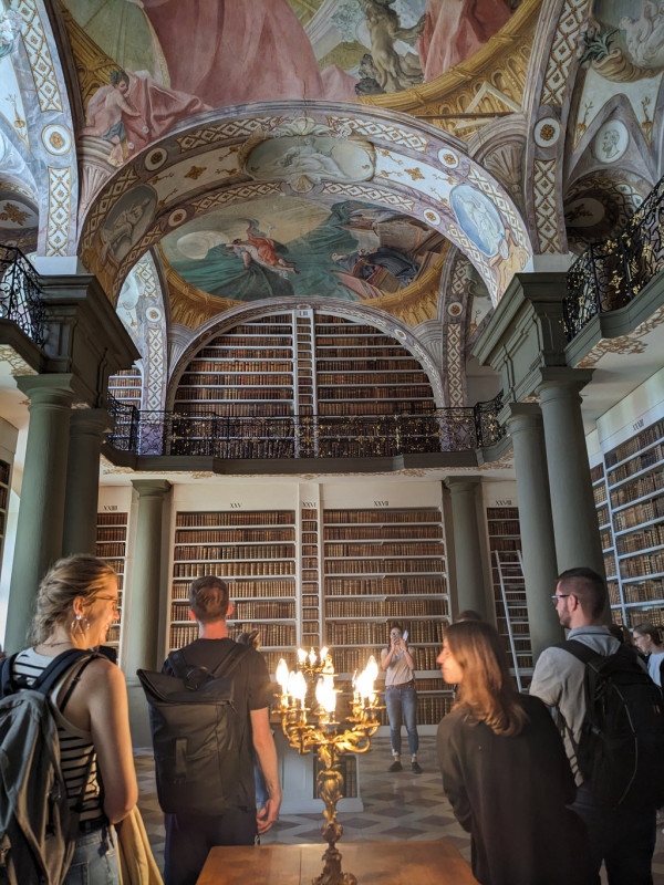 Man sieht einen prächtigen Bibliotheksraum mit Säulen, einer Galerie und Deckenfresken auf einer gewölbten Decke. Im Vordergrund sind Personen zu sehen.