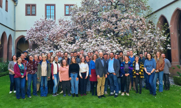 Eine Gruppe Menschen steht im Innenhof eines Gebäudes auf einer Wiese. Im Hintergrund steht ein blühender Magnolienbaum