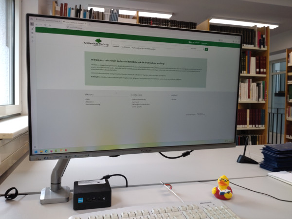 Man sieht den Bildschirm eines Bibliotheksrechners mit der Einstiegsseite des neuen Katalogs, rechts daneben eine gelbe Plastikente, die kürzlich das Maskottchen eines unserer Lehrgänge war.