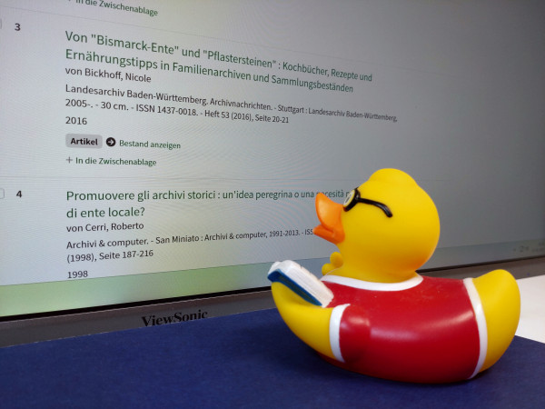 Man sieht einen Bildschirmausschnitt mit einer Trefferliste aus dem neuen Katalog, direkt davor die gelbe Plastikente, die auf den Eintrag mit dem Titel „Von Bismarck-Ente und  Pflastersteinen“ blickt.